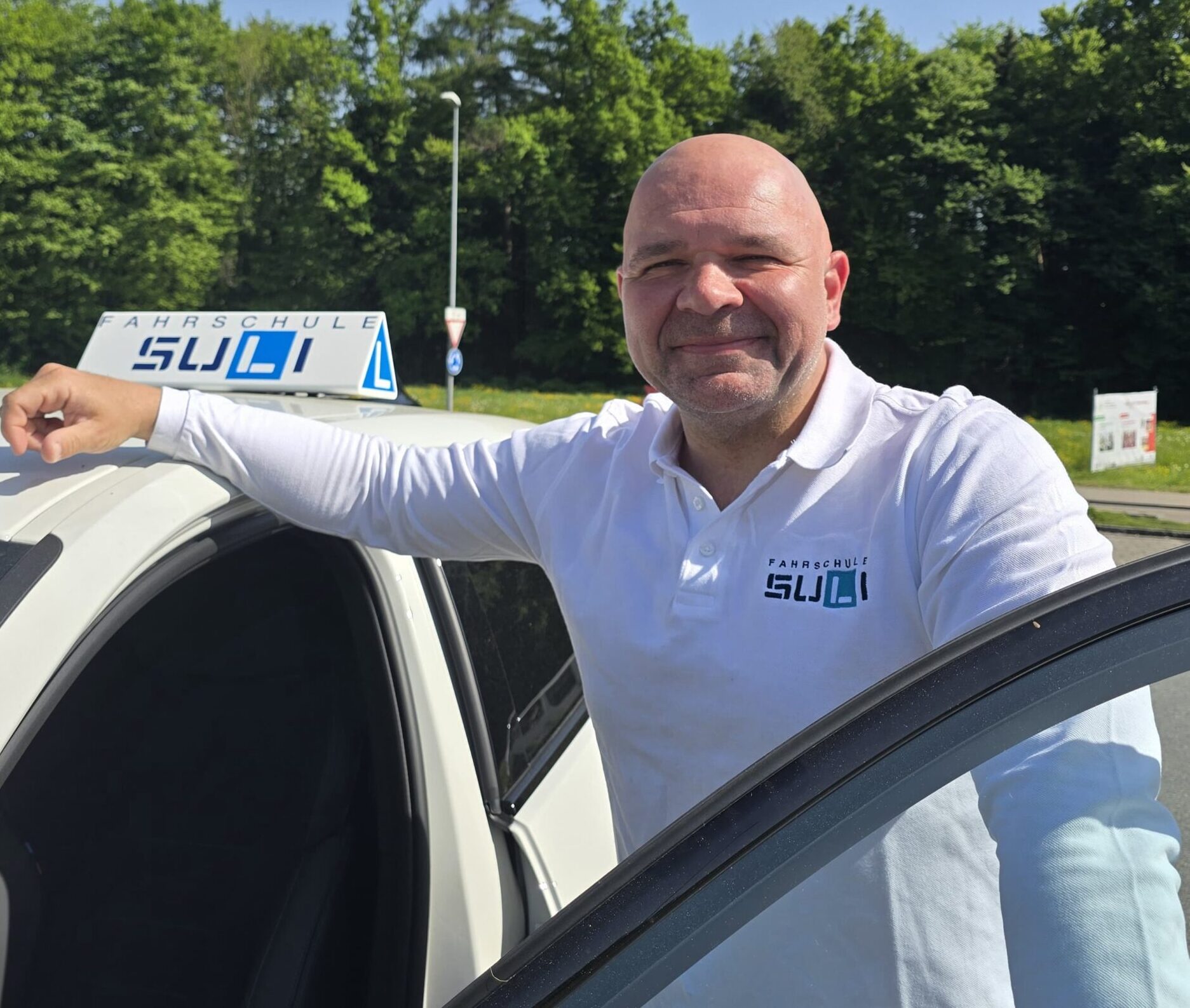Fahrlehrer Kris von der Fahrschule SULI steht lächelnd vor einem Fahrschulauto.