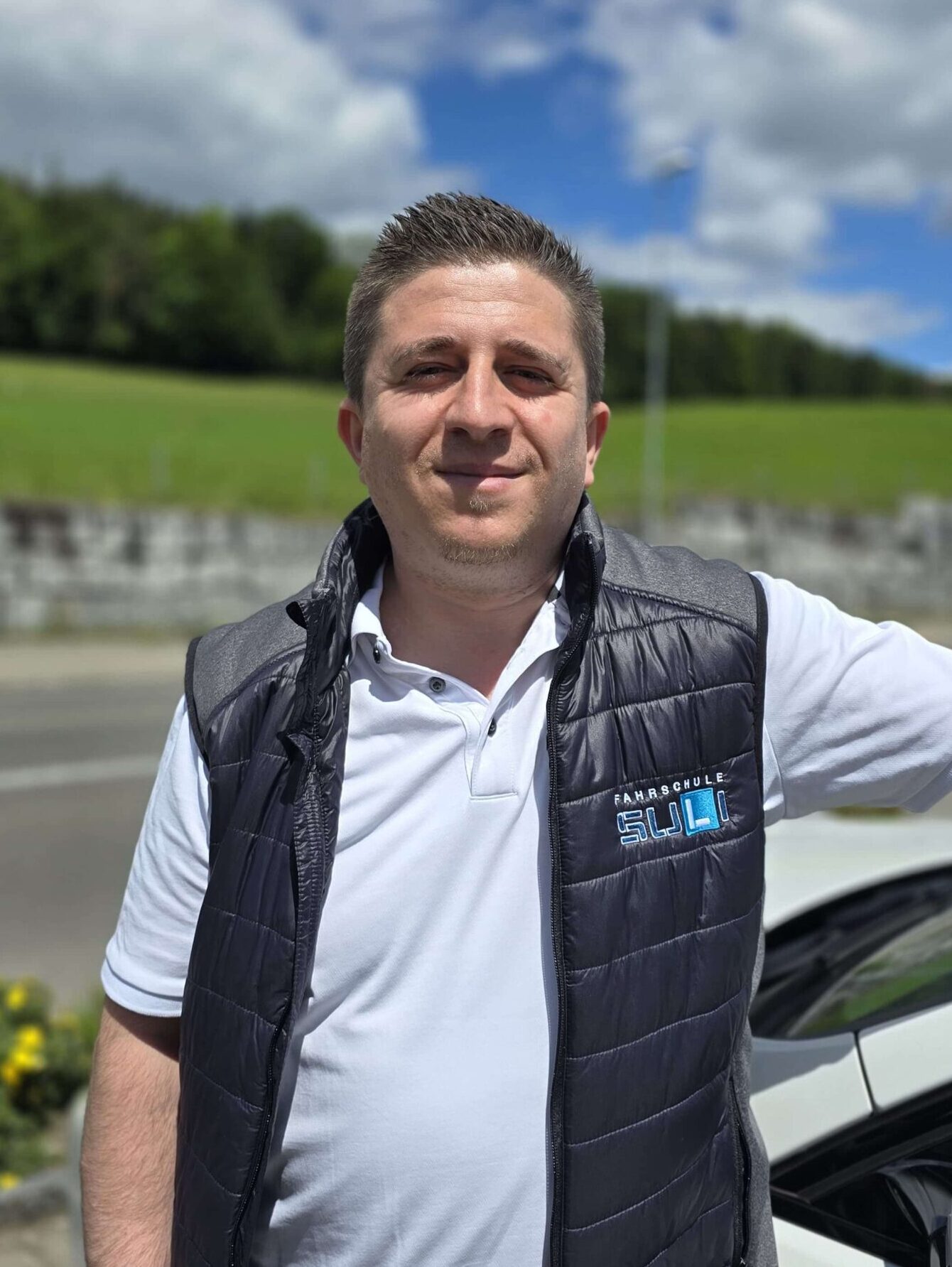 Fahrlehrer Adnan von der Fahrschule SULi steht vor einem Auto und lächelt in die Kamera.