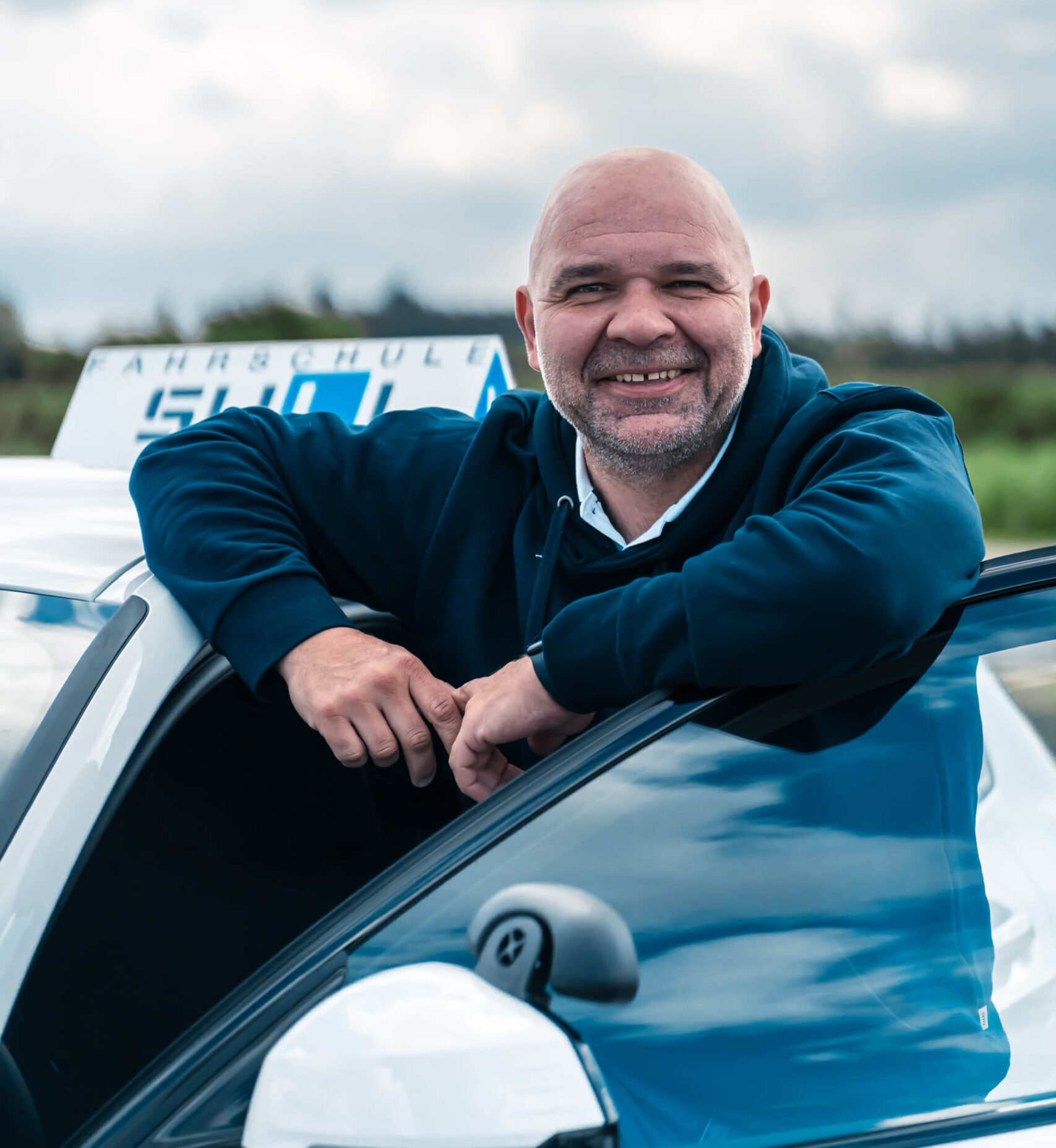 Fahrlehrer Kris von der Fahrschule SULI steht lächelnd vor einem Fahrschulauto.