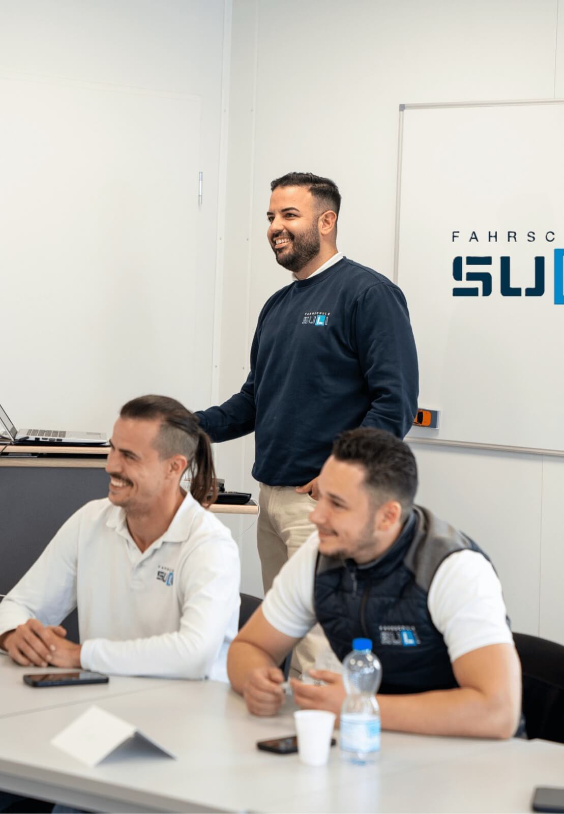 Mehrere junge Fahrschüler sitzen in einem freundlichen Kursraum der Fahrschule SULI und verfolgen aufmerksam den VKU-Unterricht, während der Fahrlehrer vorne am Whiteboard eine Verkehrssituation erklärt.