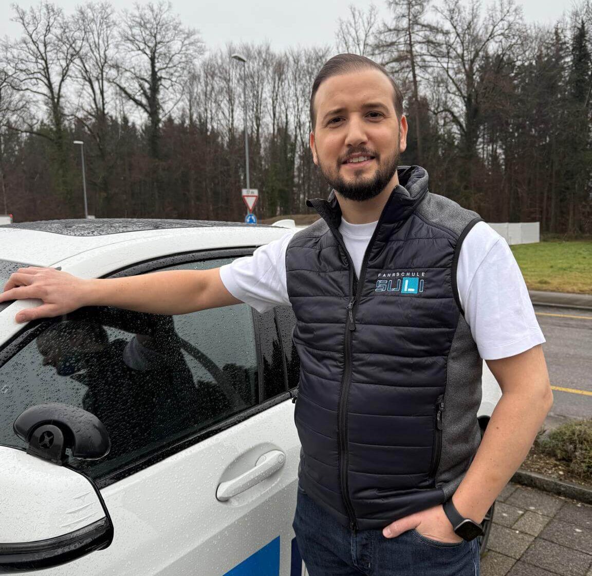 Ege, Fahrlehrer bei der Fahrschule SULI im Zürcher Oberland, steht freundlich neben dem Fahrschulauto.
